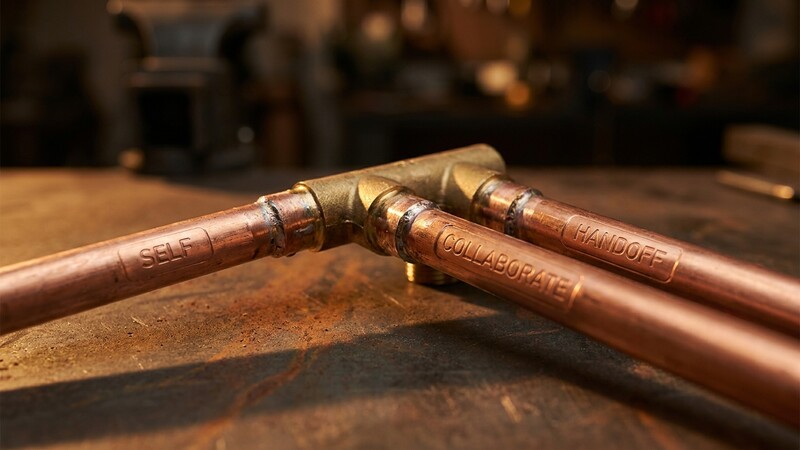 Warm amber-lit close-up of three diverging copper pipe junctions soldered onto a central brass manifold, each pipe stamped with shallow embossed labels SELF, COLLABORATE, and HANDOFF, resting on a dark oxidized steel workbench